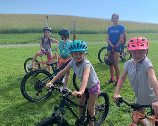 A group of young mountain bikers wearing helmets, gathered with their bikes in a grassy area.